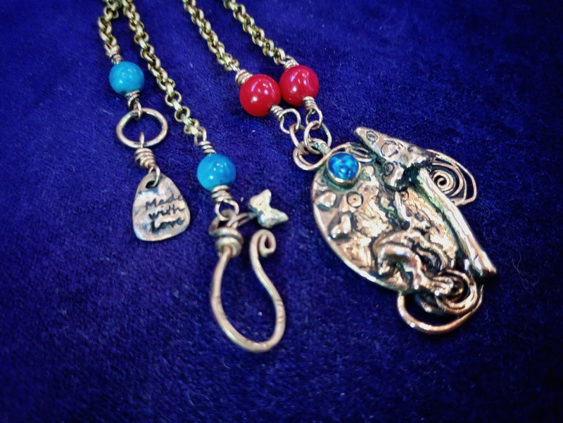 A handmade bohemian-style necklace featuring a large silver-toned paisley pendant with turquoise accents, alongside two smaller dangling charms on gold-toned chains with red and turquoise beads, displayed on a dark blue velvet background.