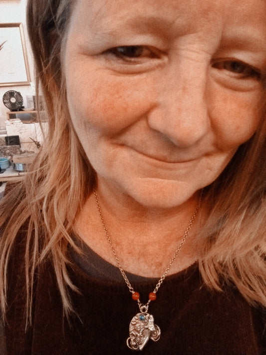 A close-up selfie of a woman wearing a silver-toned necklace with a small elephant pendant and red beads, in an indoor setting.