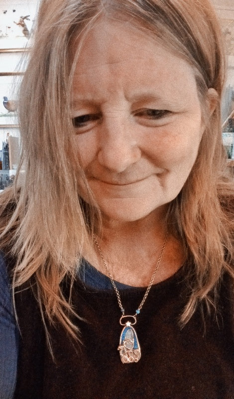 A close-up shot of a woman with light brown hair wearing a gold pendant necklace with a blue and white enamel design.