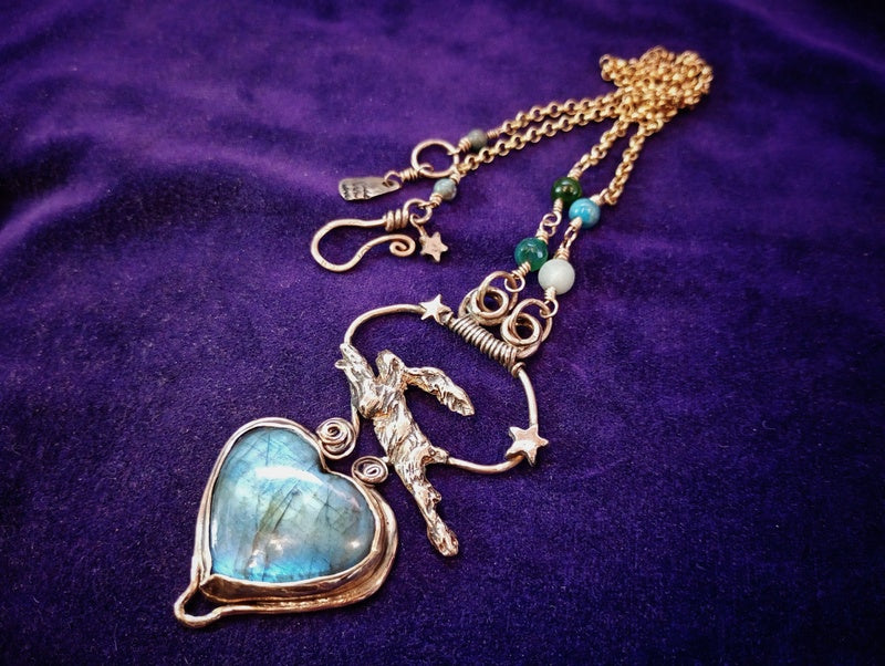 A handmade-style pendant necklace featuring a large heart-shaped labradorite stone with a blue sheen, a silver-toned unicorn charm, star details, and a gold-toned chain with small beads, displayed on a textured purple fabric background.
