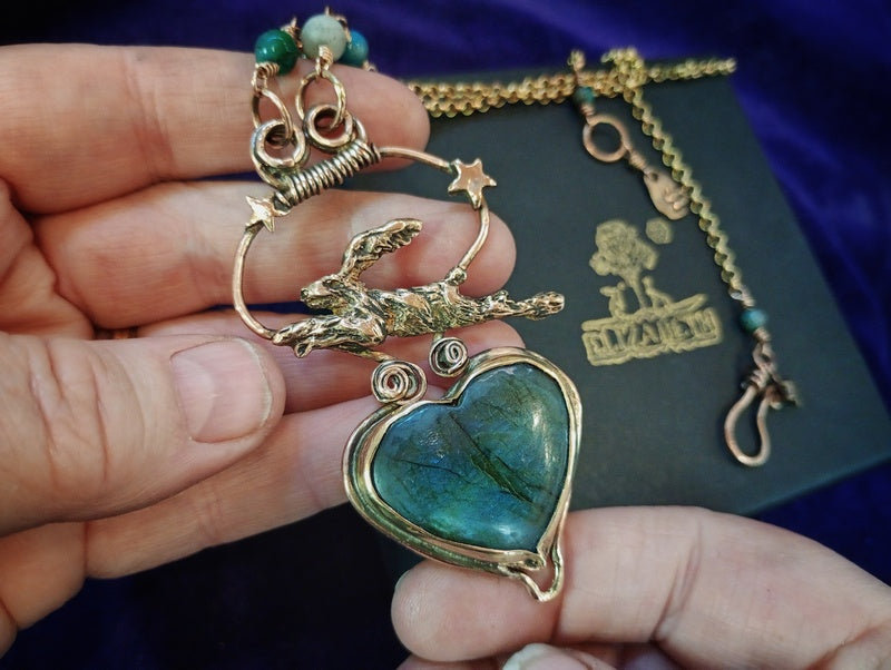 A handmade gold-toned pendant necklace featuring a running dog charm and a heart-shaped labradorite stone, held in a person's hand against a dark purple background with a branded box.