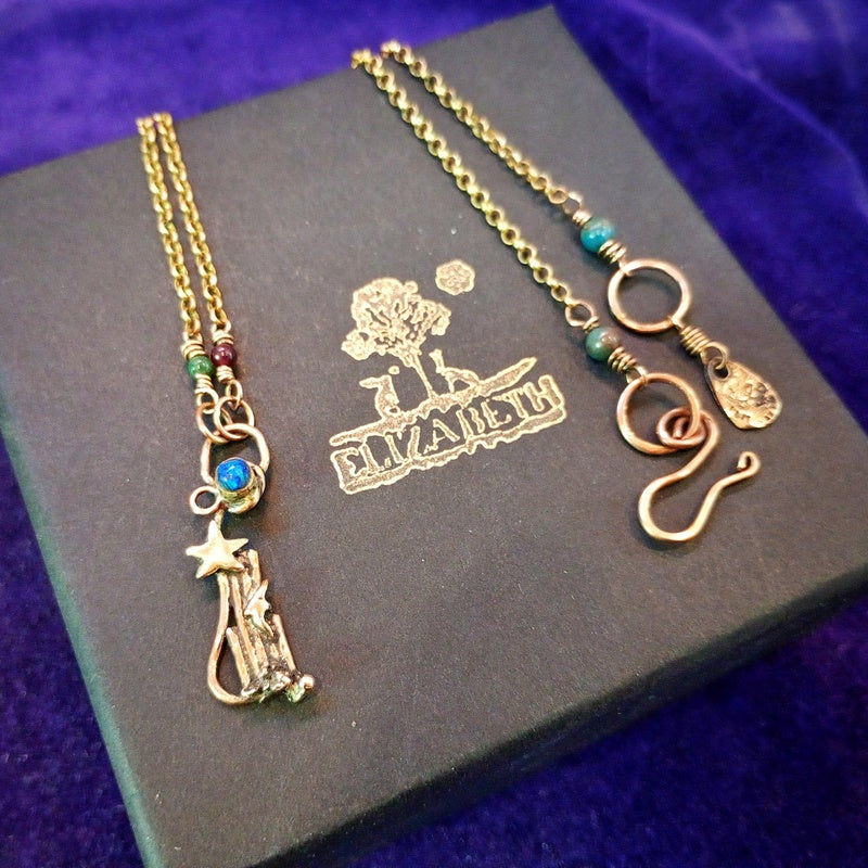 Two gold-toned jewelry pieces displayed on a dark box against a purple velvet background. One piece has a crescent moon and star pendant with small turquoise and red beads, and the other has a coiled snake charm with turquoise beads and a small tag.