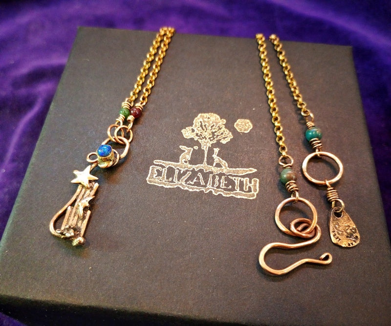 Two handmade gold-toned wire jewelry necklaces displayed on a dark brown presentation box with the brand name 'ELIZABETH' printed on it, set against a purple velvet background.