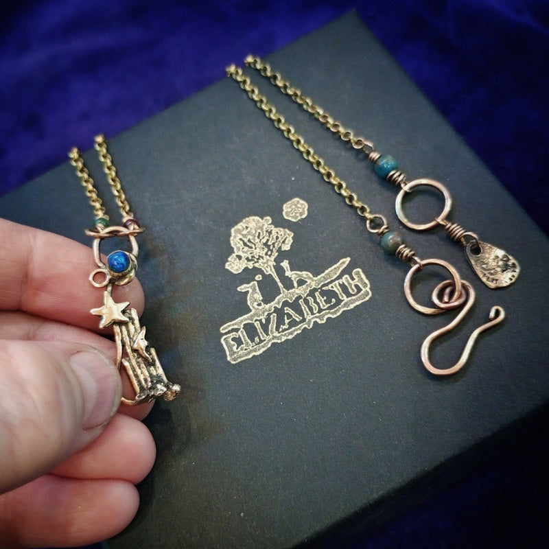 A close-up of a hand holding a gold-toned necklace with a star and basket pendant, next to another gold-toned chain necklace with turquoise beads and a teardrop charm, laid on a dark box with a gold 'ELIZABETH' logo, against a deep blue velvet background.