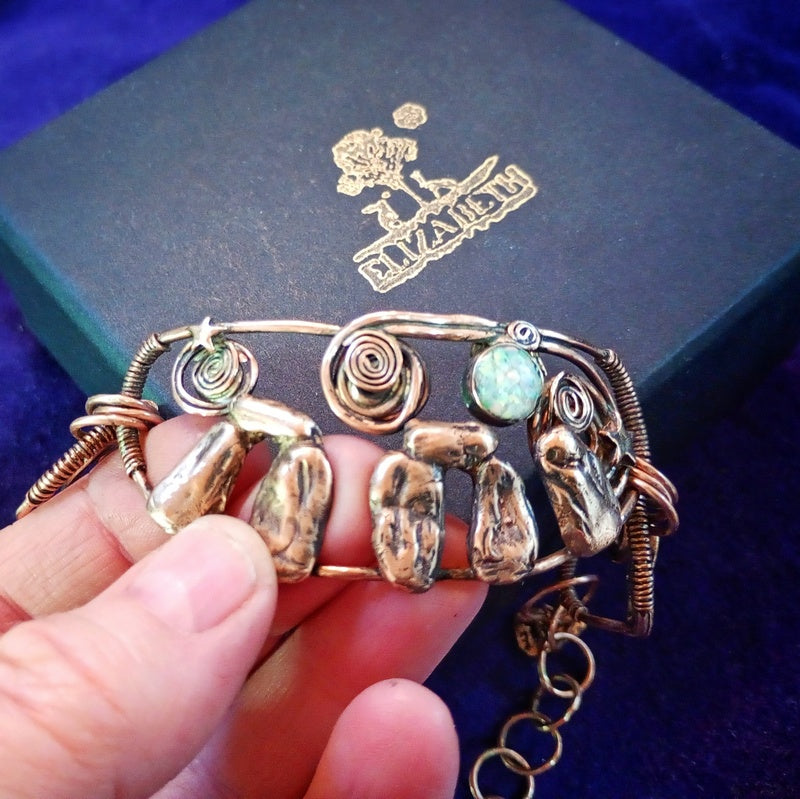A handmade wire-wrapped bracelet with textured metallic charms and a round turquoise stone, held by a hand in front of a dark blue jewelry box with a gold logo. Stonehenge Bracelet.