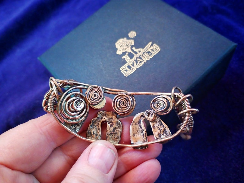 A handmade copper wire bracelet with spiral and textured details, held in a hand against a dark blue velvet background with a branded box.