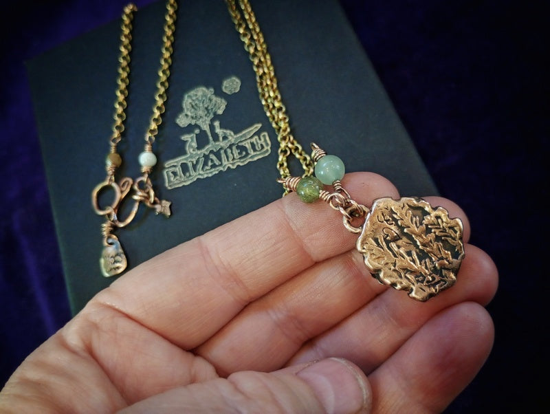A gold-toned necklace with a textured round pendant and small green beads, held in a person's hand over a dark branded box.