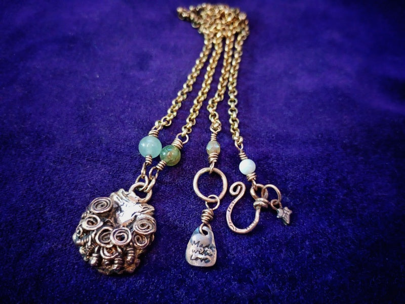 A bohemian-style multi-pendant necklace with a gold chain, featuring a large silver-toned skull pendant, a small silver-toned bird charm, a silver-toned fish charm, a gold hook charm, and light green and white beads on a dark purple velvet background.