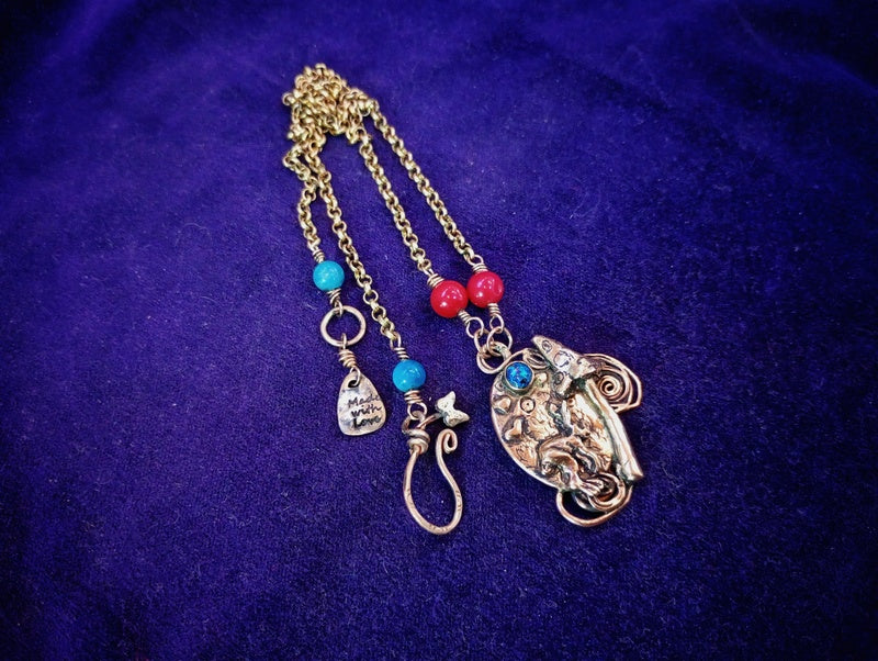 A bohemian-style gold-toned chain necklace featuring a large elephant pendant, turquoise and red beads, and small charm details, displayed on a textured purple fabric background.