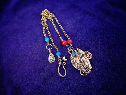 A bohemian-style gold-toned chain necklace featuring a large elephant pendant, turquoise and red beads, and small charm details, displayed on a textured purple fabric background.