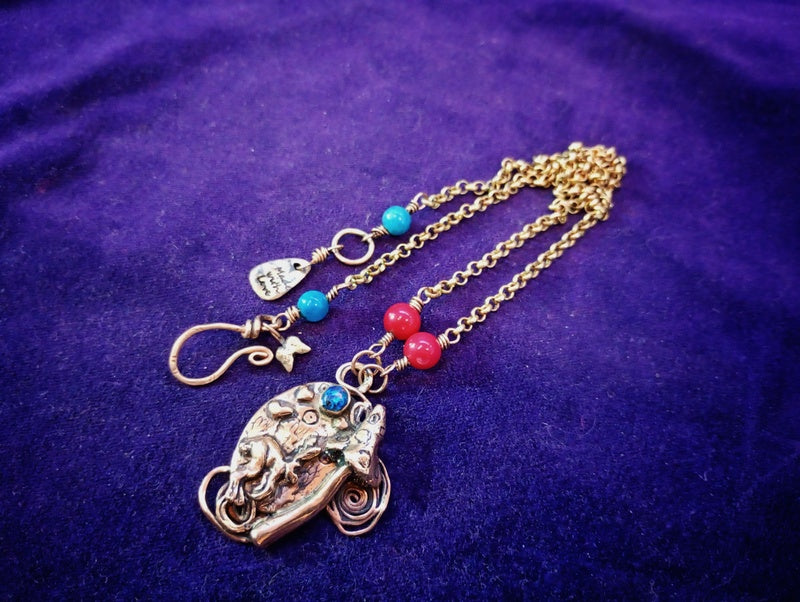 A handmade bohemian-style necklace featuring a detailed elephant pendant, a gold chain, and small turquoise and red beads, displayed on a textured purple fabric background.