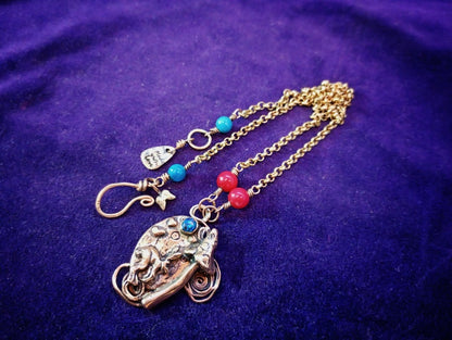 A handmade bohemian-style necklace featuring a detailed elephant pendant, a gold chain, and small turquoise and red beads, displayed on a textured purple fabric background.