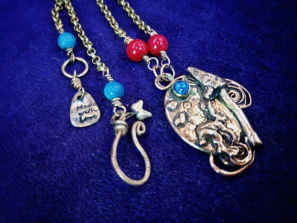 A handmade bohemian-style necklace featuring a large silver-toned paisley pendant with turquoise accents, alongside two smaller dangling charms on gold-toned chains with red and turquoise beads, displayed on a dark blue velvet background.