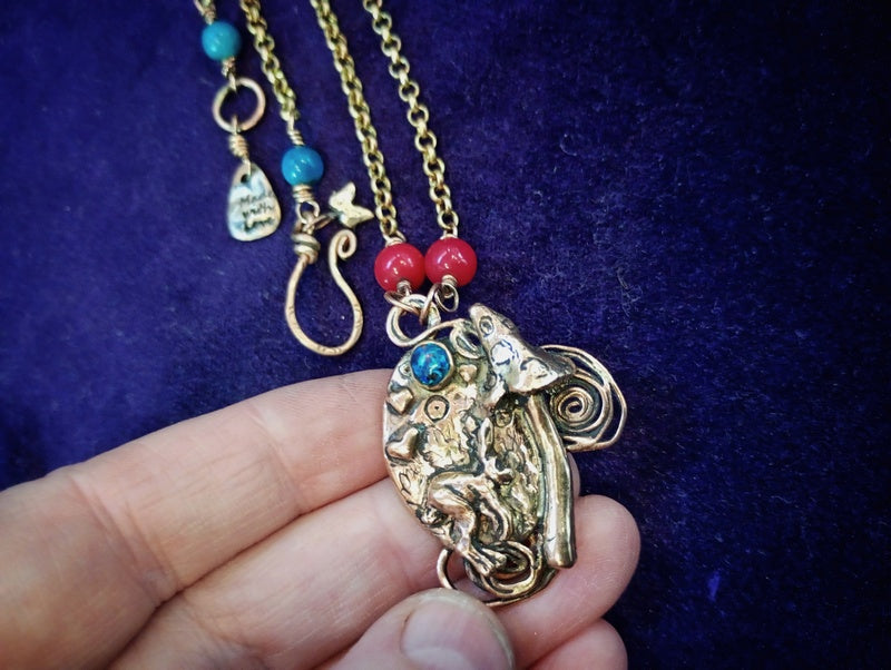 A hand holding a gold-toned pendant necklace featuring a detailed frog design with turquoise and red bead accents, set against a dark purple velvet background.