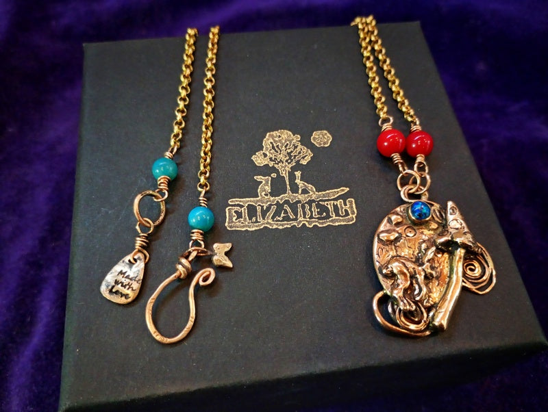 Two gold-toned jewelry necklaces displayed on a black box with the brand name Elizabeth. One necklace has turquoise beads and a small pendant, while the other has red beads and a large ornate elephant-shaped pendant with a blue accent.