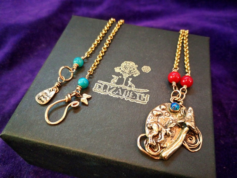 A gold-toned necklace with a detailed elephant pendant, turquoise and red beads, and a branded black box on a purple fabric background.