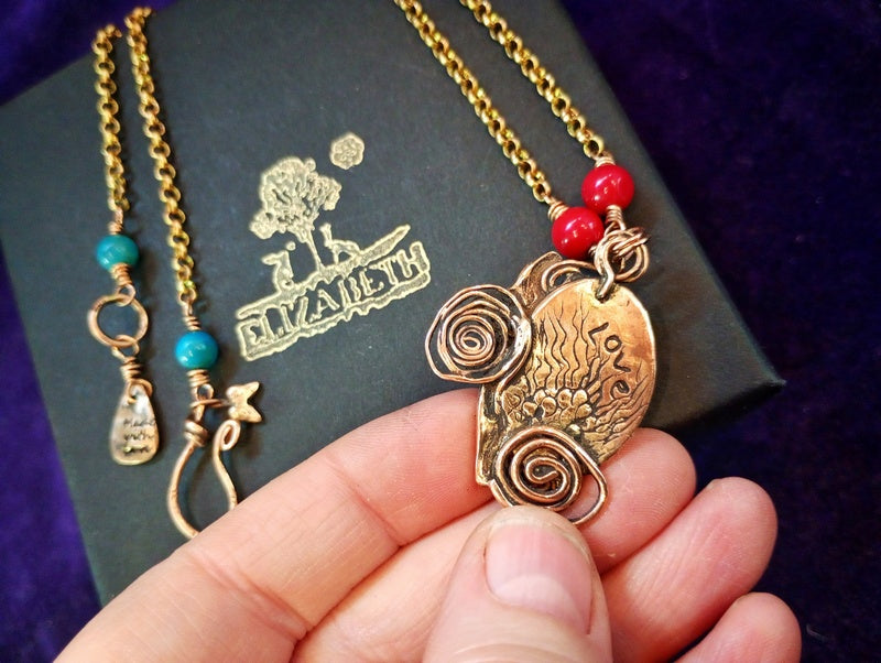 A close-up of a hand holding a gold-toned pendant necklace with red beads, with another necklace and a dark branded box in the background.