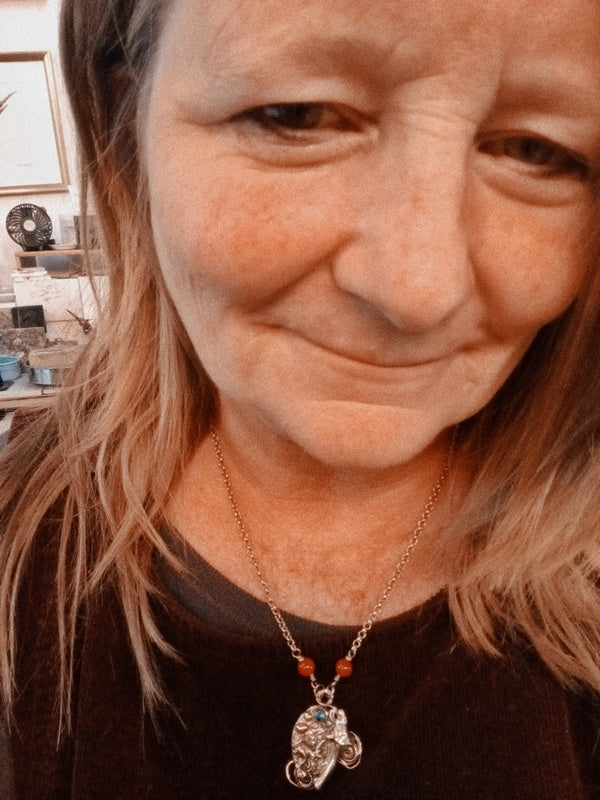 A close-up selfie of a woman wearing a silver-toned necklace with a small elephant pendant and red beads, in an indoor setting.