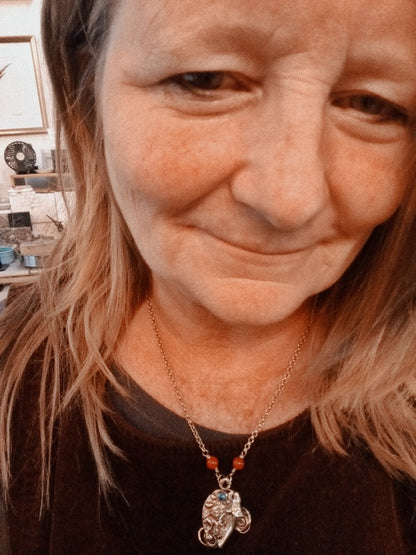 A close-up selfie of a woman wearing a silver-toned necklace with a small elephant pendant and red beads, in an indoor setting.