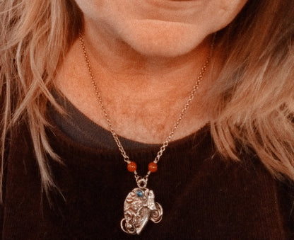 A close-up of a person wearing a silver-toned elephant pendant necklace with small orange beads and a blue stone accent, shown against a dark brown top.
