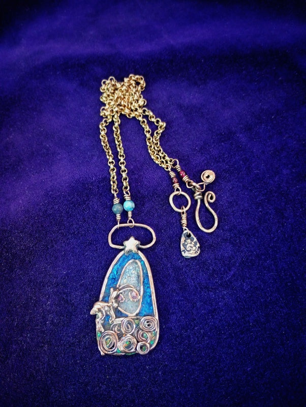 A handmade gold chain necklace featuring a blue and silver filigree pendant with a small turquoise bead and a dangling clear teardrop charm, displayed on a dark purple velvet background.