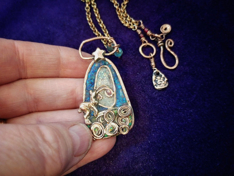 A handmade gold pendant necklace with turquoise blue enamel and wire-wrapped details, held by a hand against a dark purple velvet background.