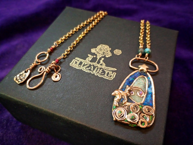 A gold-tone filigree pendant necklace with blue, green, and red enamel details, displayed on a black Elizabeth box over a purple fabric background.