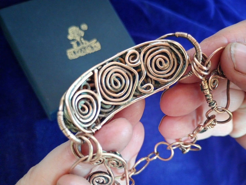 Bronze bracelet with intricate designs swirls on the back held by a hand against a blue background