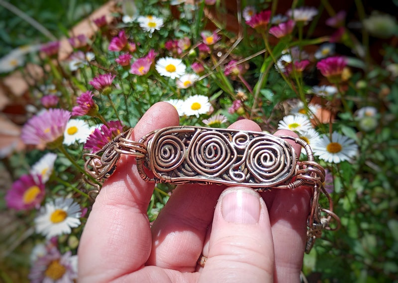 Hand holding a bronze bracelet with intricate designs against a background of flowers
