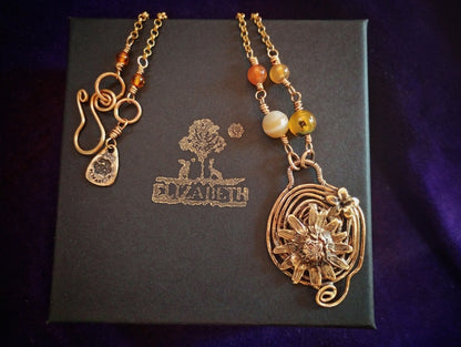 A handmade gold-toned beaded necklace with a large sunflower pendant and spiral wire details, displayed on a dark gray box with the brand name 'ELIZABETH' printed on it, set against a dark purple background.