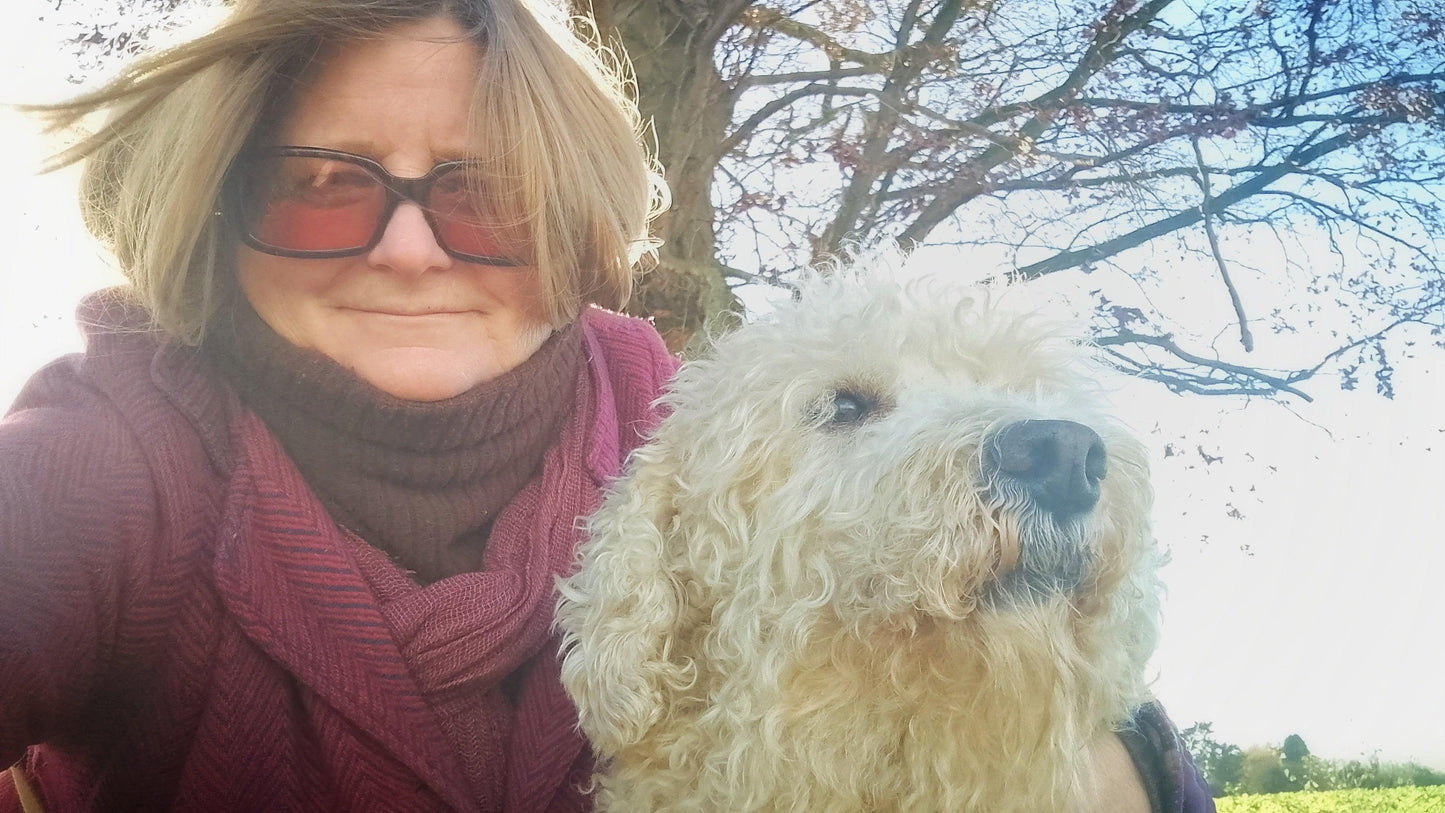 A woman wearing sunglasses, a dark turtleneck, and a burgundy cardigan holds a fluffy cream-colored dog outdoors under a tree free animal healing Elizabeth Shewan