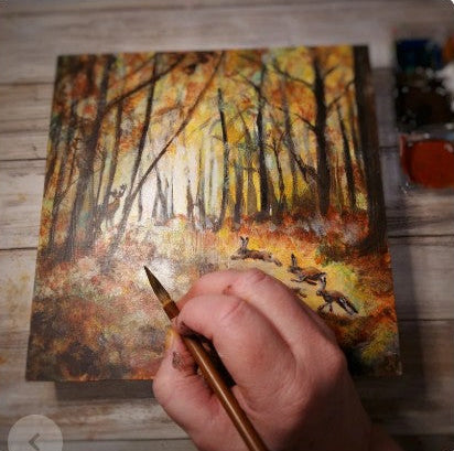 artists hand holding a paintbrush over a painting by Elizabeth Shewan of a forest scene on a wooden surface.
