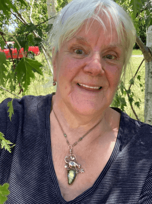 Woman with short white hair wearing a dark top and necklace with a green labradorite pendant made by artist and jewellery designer Elizabeth Shewan standing outdoors with trees and a red vehicle in the background.