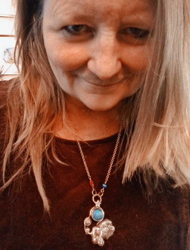 A close-up lifestyle shot of a woman with blonde hair wearing a silver-toned necklace with a small elephant pendant and a round blue stone accent.
