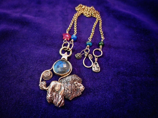 A handmade gold-toned pendant necklace featuring a lion and dog charm, a blue stone, and small colorful beads, displayed on a purple velvet background.