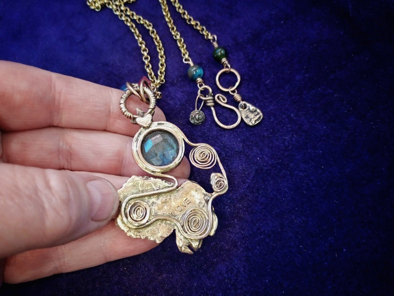 A handmade gold-toned pendant necklace with a blue-green labradorite stone and spiral wire details, held by a hand against a dark blue velvet background.