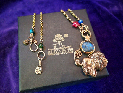 A gold-toned statement necklace with a lion head pendant, blue stone, and colorful beads, displayed on a black branded box against a purple velvet background.
