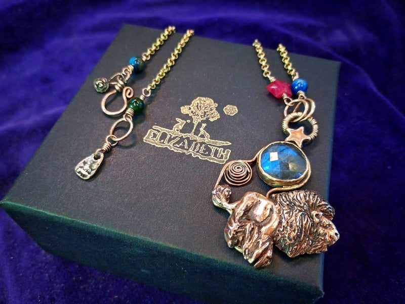 A gold-toned lion pendant necklace with a blue faceted gemstone and colorful beads, displayed on a dark green jewelry box with the brand name 'Dizaji' printed on it, set against a purple velvet background.