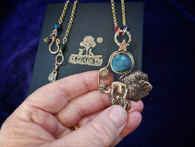 A hand holding a gold-tone pendant necklace featuring a lion figurine, a star, and a round blue stone, with another necklace and a branded black display card in the background.