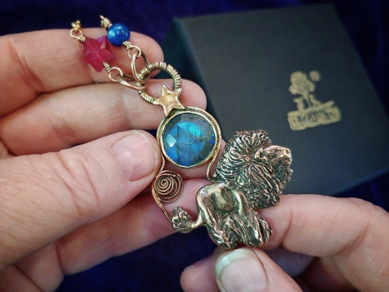A close-up of a hand holding a gold-toned lion pendant necklace with a blue labradorite stone and red and blue beads, with a black presentation box in the background.