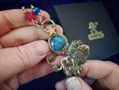 A close-up of a hand holding a gold-toned lion pendant necklace with a blue labradorite stone and red and blue beads, with a black presentation box in the background.