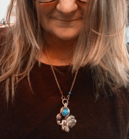 A close-up lifestyle shot of a woman wearing a silver-toned necklace with a silver elephant pendant and a round turquoise blue stone, paired with a dark brown top.