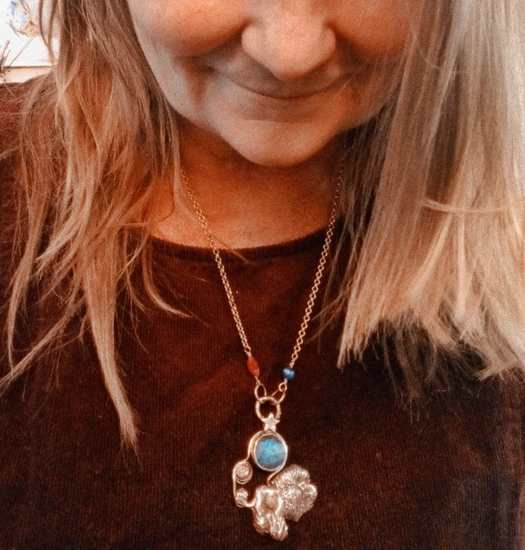 A close-up lifestyle shot of a woman wearing a silver-toned pendant necklace with a blue stone and small orange and blue beads on the chain, paired with a brown knit top.