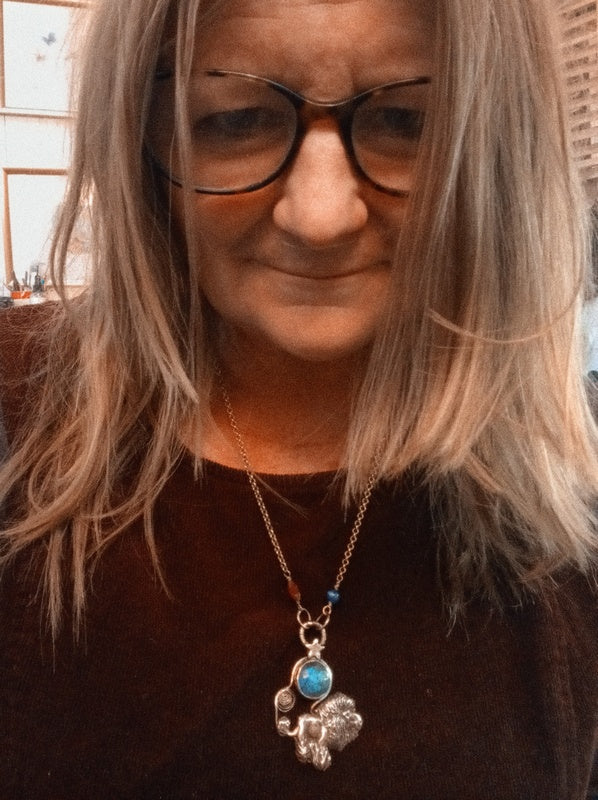 A close-up shot of a woman with gray hair and glasses wearing a silver-toned necklace with a silver elephant pendant and a round turquoise blue stone.