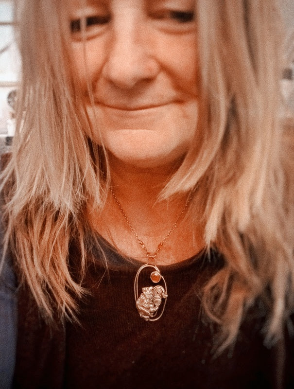 A close-up lifestyle shot of a woman with blonde hair wearing a delicate gold necklace with a unique oval pendant featuring a textured gold design and a small orange accent stone.