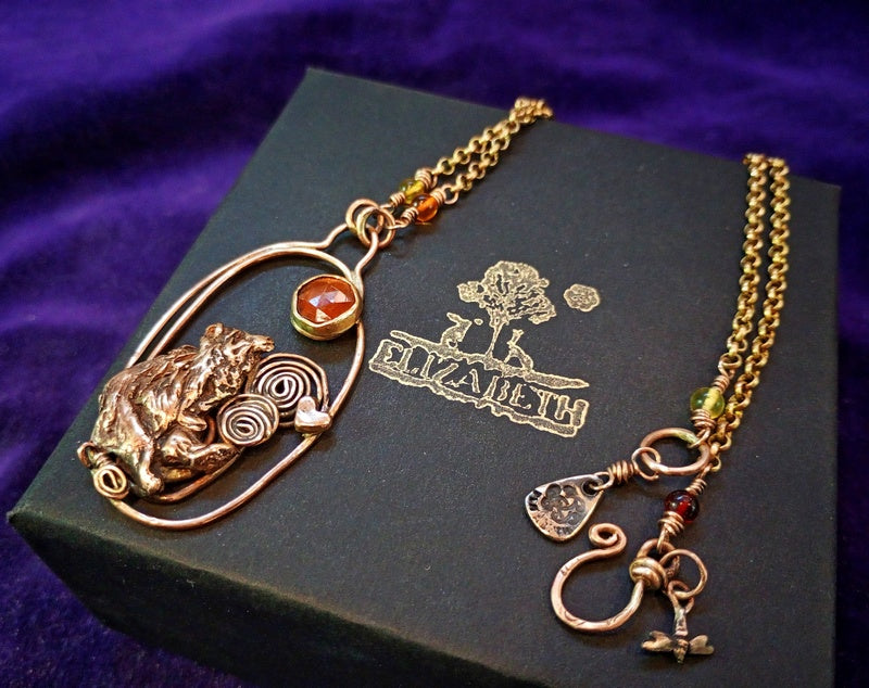 A gothic-style Celtic knot necklace with a toad pendant and amber gemstone, displayed on a black jewelry box with 'Elizabet' printed on it, set against a purple fabric background.