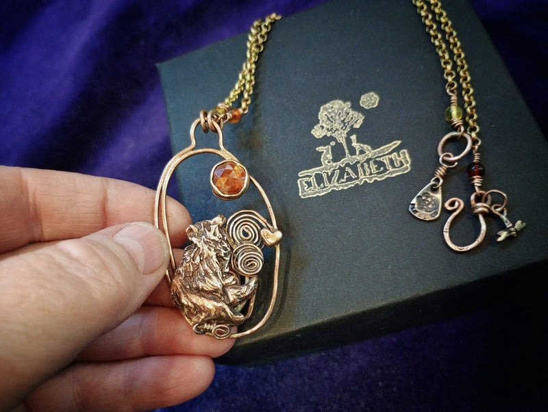 A hand holds a bronze-toned pendant necklace featuring a frog and spiral design with a small amber stone, next to a dark box branded 'DIVA ART' with a matching chain and charms laid out on a purple fabric background.