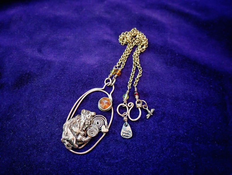 A handmade bohemian-style pendant necklace featuring a silver-toned textured animal head, a gold-toned oval frame, a small amber stone, and multiple dangling charms including a crescent moon, a teardrop charm, and a cross, displayed on a deep purple velvet background.