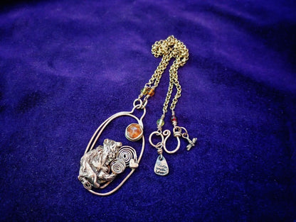 A handmade bohemian-style pendant necklace featuring a silver-toned textured animal head, a gold-toned oval frame, a small amber stone, and multiple dangling charms including a crescent moon, a teardrop charm, and a cross, displayed on a deep purple velvet background.