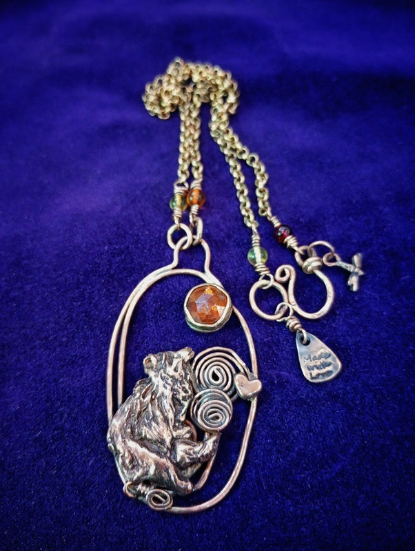 A handmade pendant necklace featuring a silver-toned bear figurine, a spiral motif, a small heart, and a faceted orange stone, displayed on a deep purple velvet background.